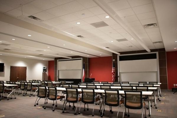 Large room with several rows of white tables and black chairs facing two projector screens on a red wall with a podium in the center.